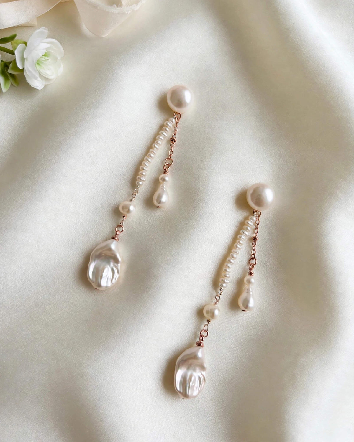 A pair of dangle earrings with large white pearls, small rose gold chain links, and teardrop-shaped clear shell charms, displayed on a soft cream fabric background.