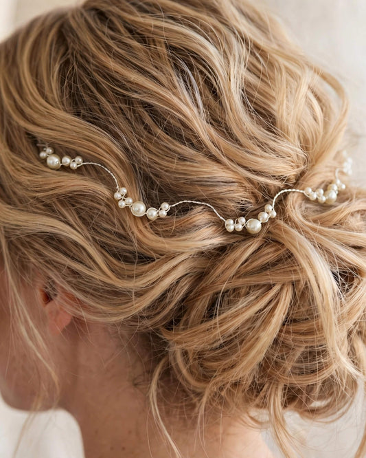 A close-up of a woman's styled blonde updo featuring a delicate silver wire hair accessory adorned with small white pearls, shown from the side.