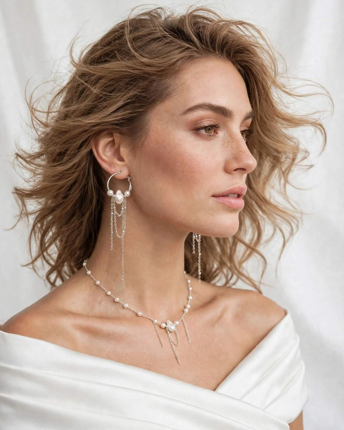 A woman in a white off-the-shoulder top wearing delicate silver-toned jewelry, including a beaded necklace, matching drop earrings, and a pearl-and-silver headpiece, against a soft white draped background.